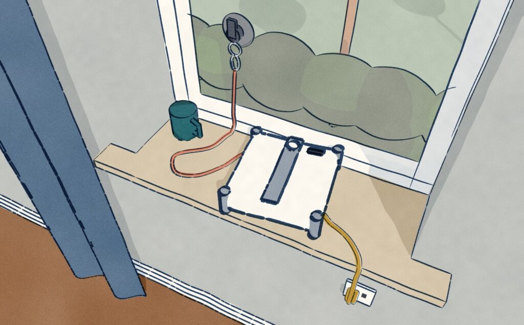 Close on an upstairs window sill, a square white window-cleaning robot lies face up where it was carried and set down, its coral safety rope clipped to a suction anchor stuck on the glass above it and its gold power cord run along the sill and down into a socket under it, with the garden treetops below the window.