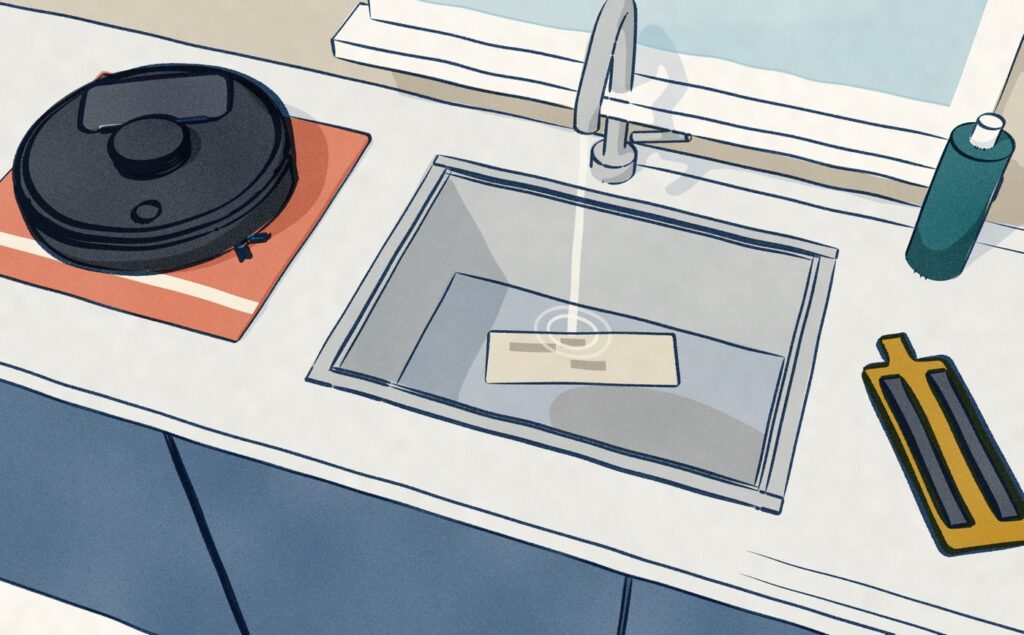 From above a kitchen worktop: the black Ecovacs Neo 4.0 sits lifted onto a coral tea towel beside the sink, its mop plate unclipped on the worktop, while its grey pad lies in the steel basin under the running tap.