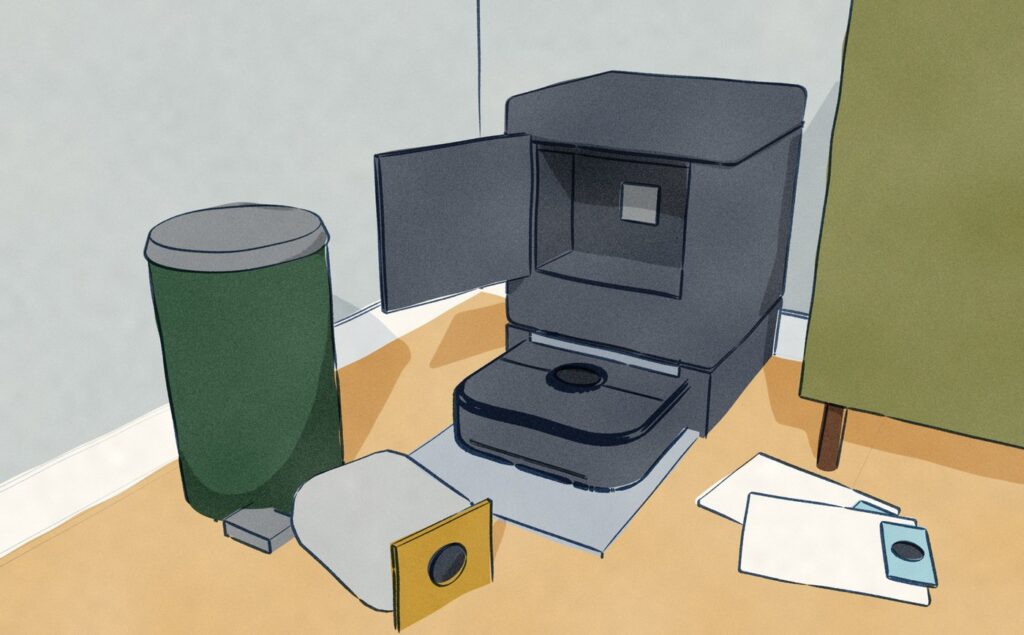 In the corner of a dining room a dark square-cornered robot vacuum sits charging in its tall dock, whose front bag door is open; the full dust bag lies on the floor beside a pedal bin and two new bags lie by the sideboard.