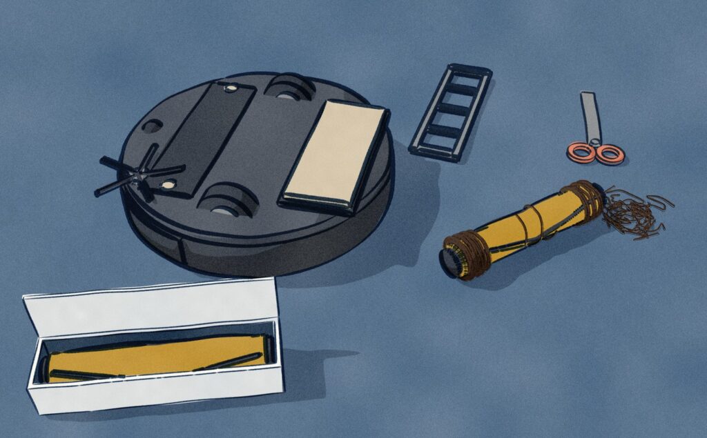 A black Roborock S7 Max Ultra lies upside down on a denim duvet at the foot of a bed, its brush guard off and its rubber brush roller lifted out with long hair wound round both ends, beside scissors, snipped hair and a new roller in its box.