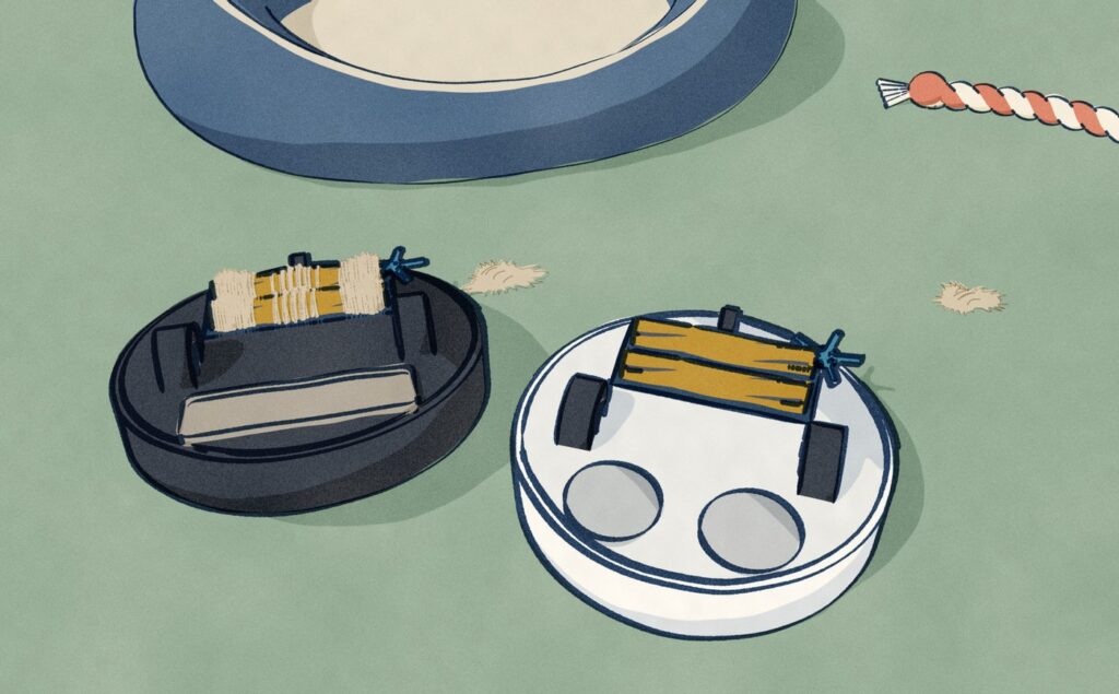 Two robot vacuums lie upside down on the carpet beside a dog bed: the black one has cream dog hair wound tight round both of its brush rollers, while the white one has bare brushes.